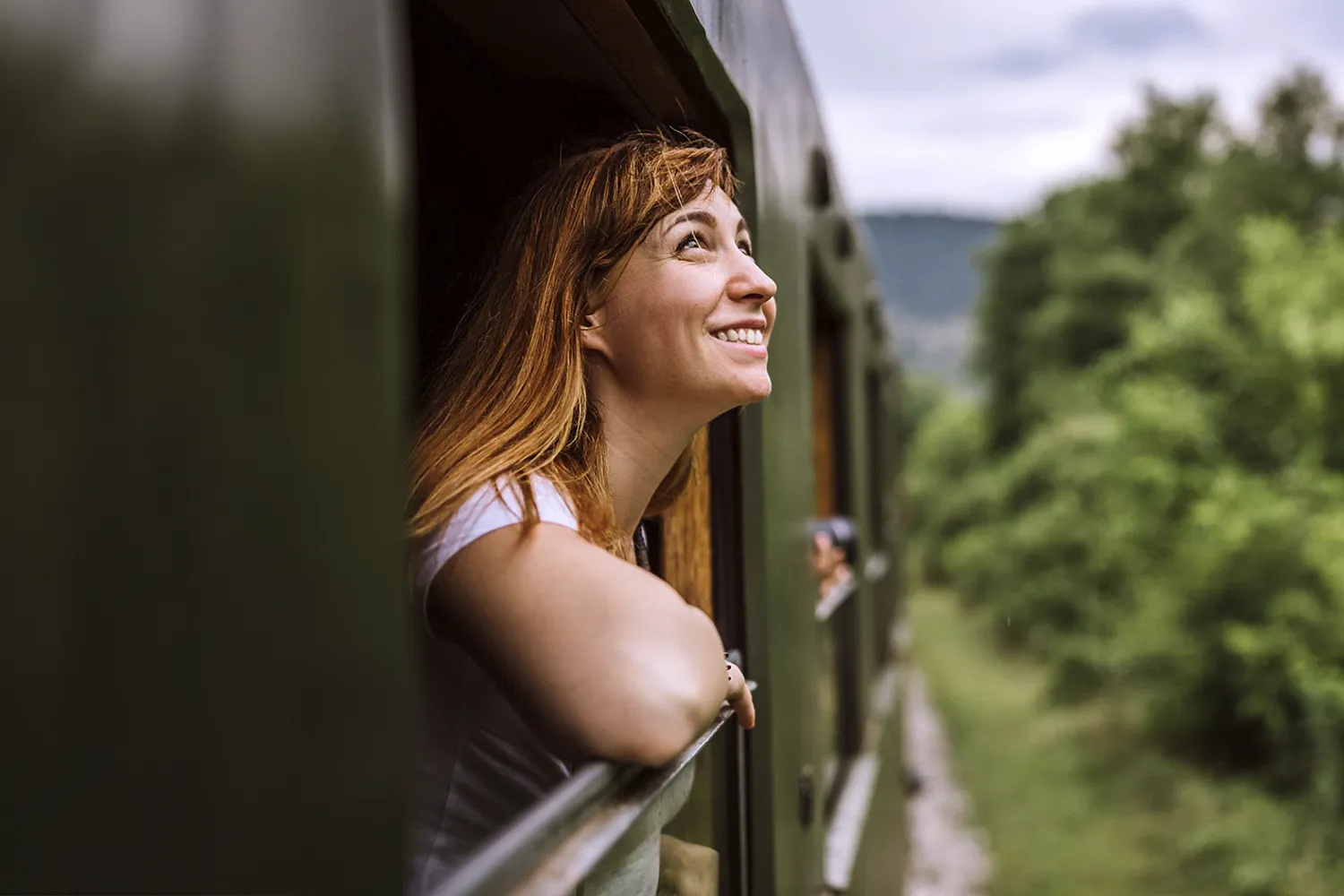 Smiling woman with red hair leaning out of a moving train window, looking up at the sky surrounded by green trees.