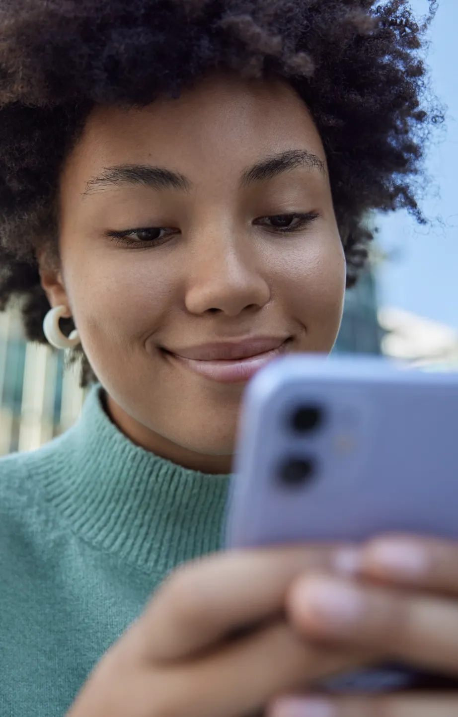 Young woman with natural hair smiling while looking at her smartphone outdoors.