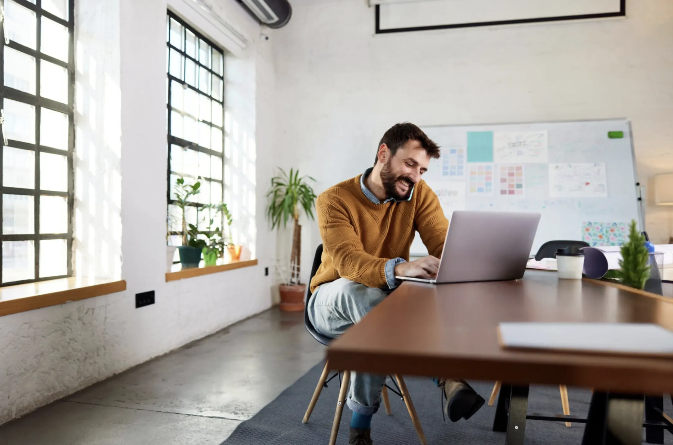 Smiling man in a mustard sweater talking on phone while working on a laptop at a desk in a bright modern office.