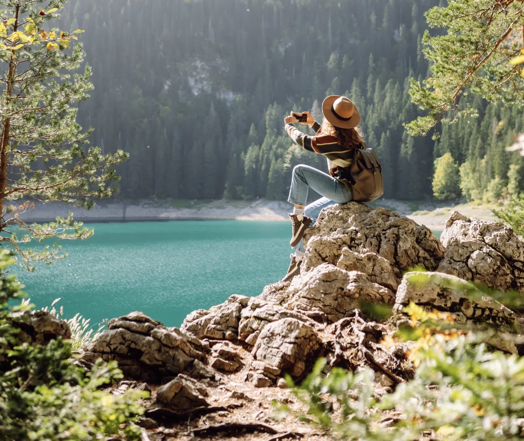 Woman in a wide-brim hat sits on a rocky outcrop photographing a turquoise mountain lake surrounded by pine forest.
