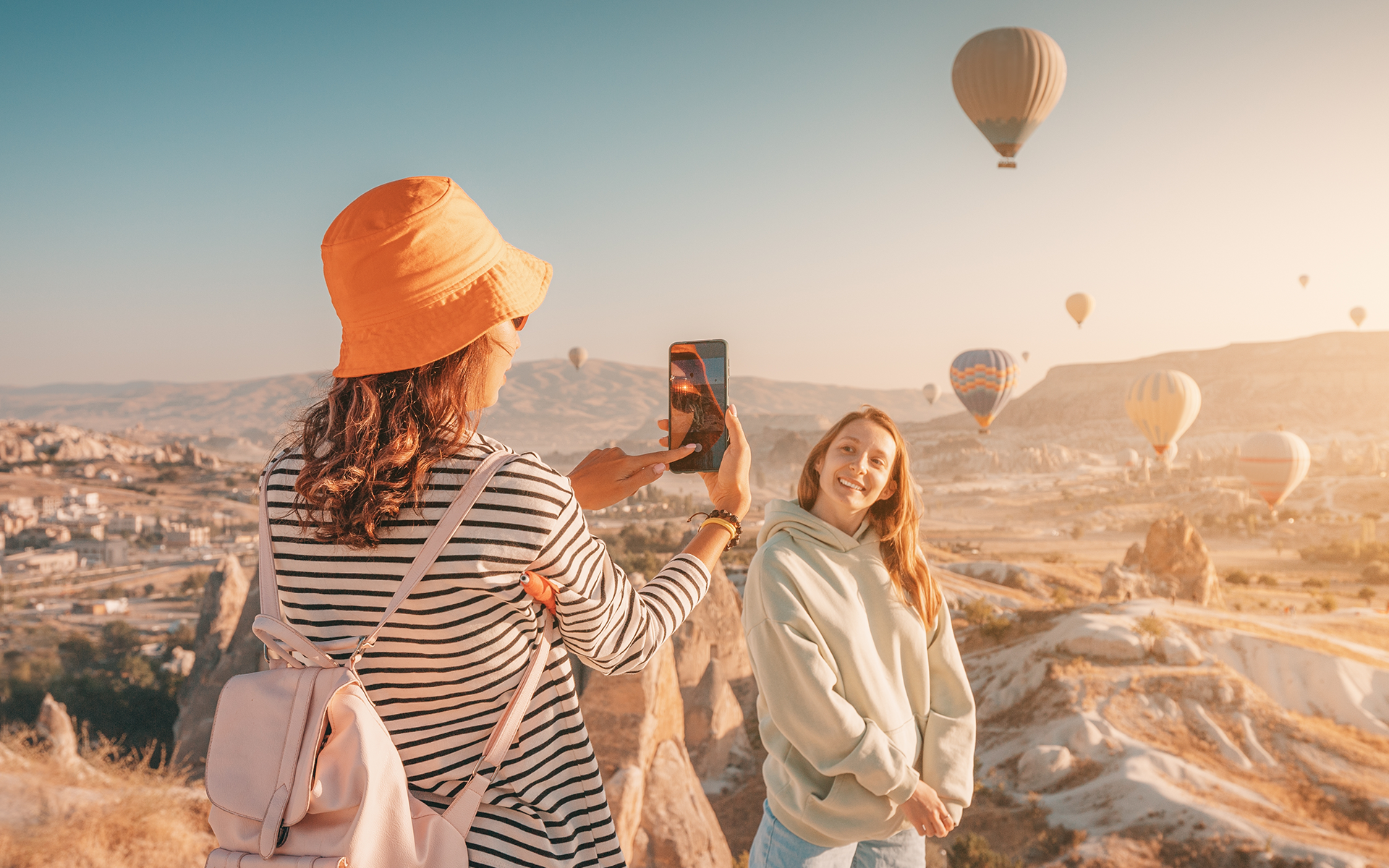 Woman in orange bucket hat photographing a smiling friend on a hilltop in Cappadocia, Turkey, with colorful hot air balloons filling the sky.