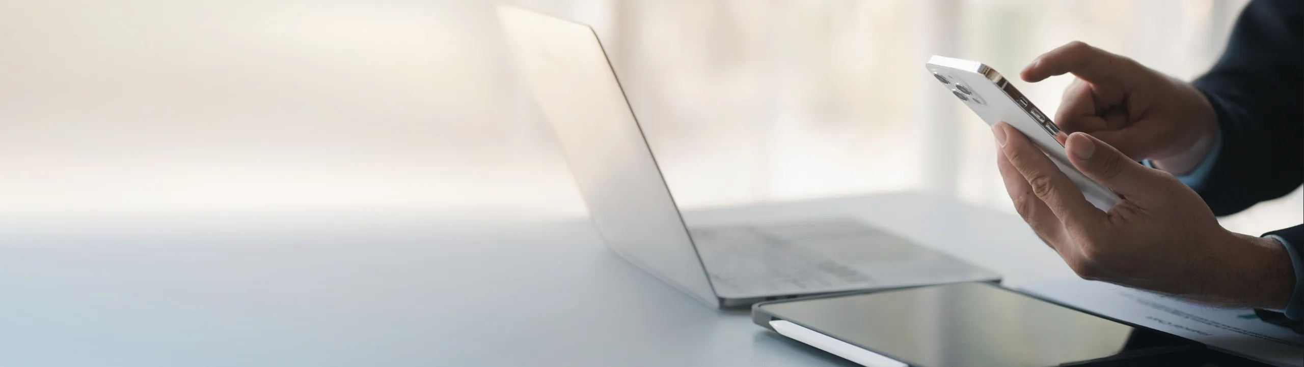 Person in a suit using a smartphone at a desk with a laptop and tablet nearby.