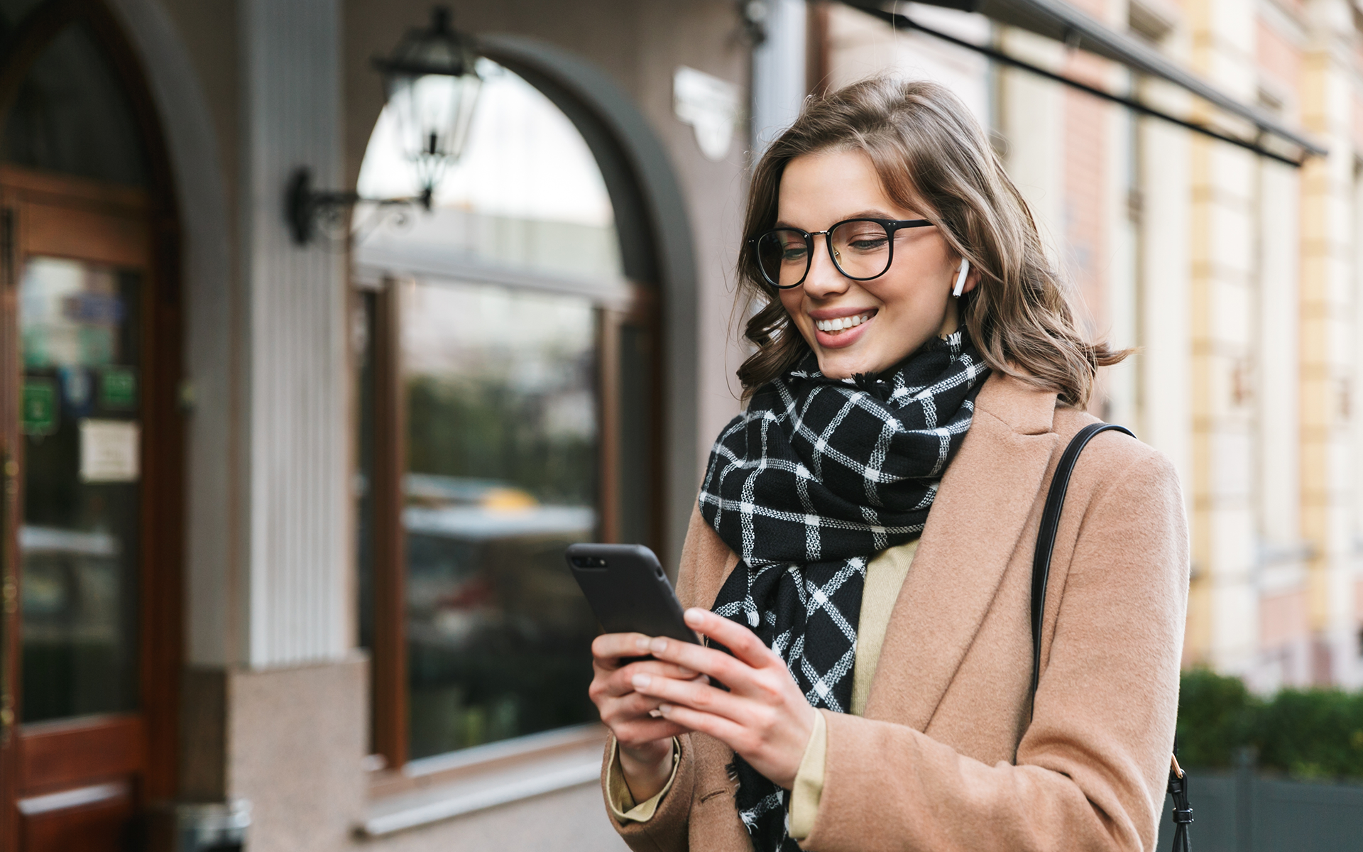 Smiling young woman with glasses and wireless earbuds using a smartphone outdoors on a city street in a tan coat and plaid scarf.