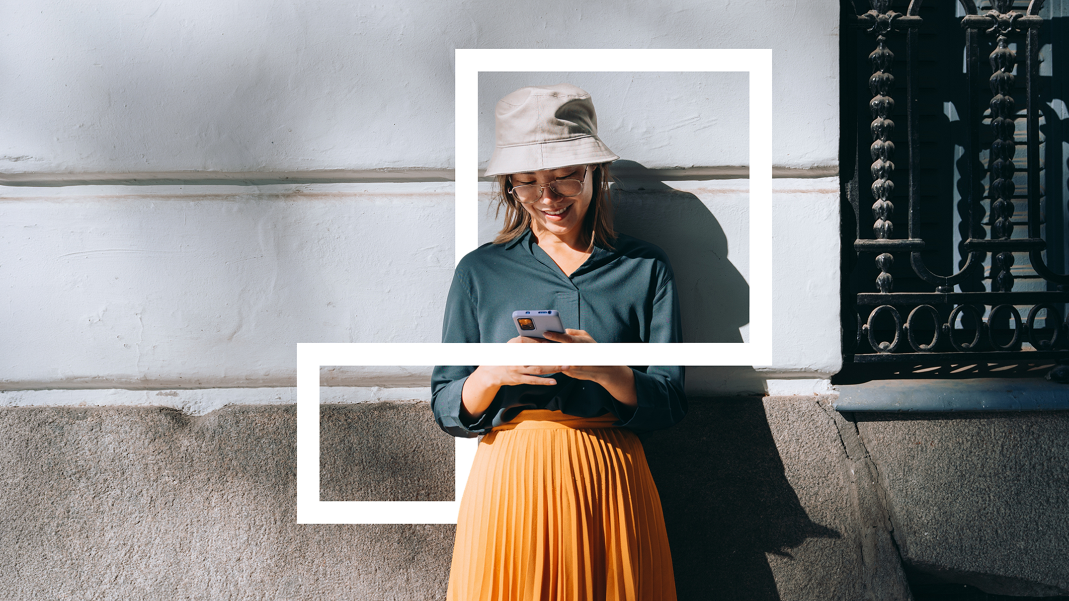 Smiling woman in a bucket hat and orange pleated skirt leaning against a wall, looking at her smartphone outdoors.