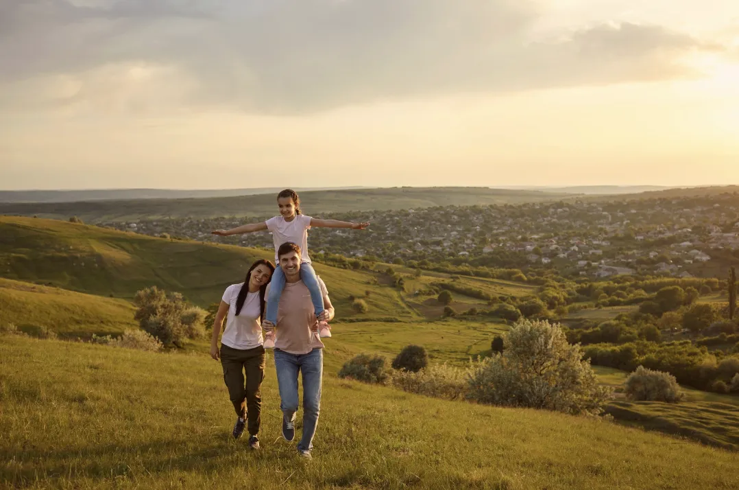 Family walking on a grassy hillside at sunset, father carrying young girl on his shoulders with arms outstretched, mother smiling beside them.