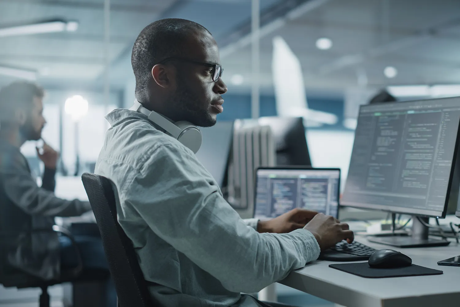 Man wearing glasses and headphones typing at a desk with dual monitors displaying code in a modern office.