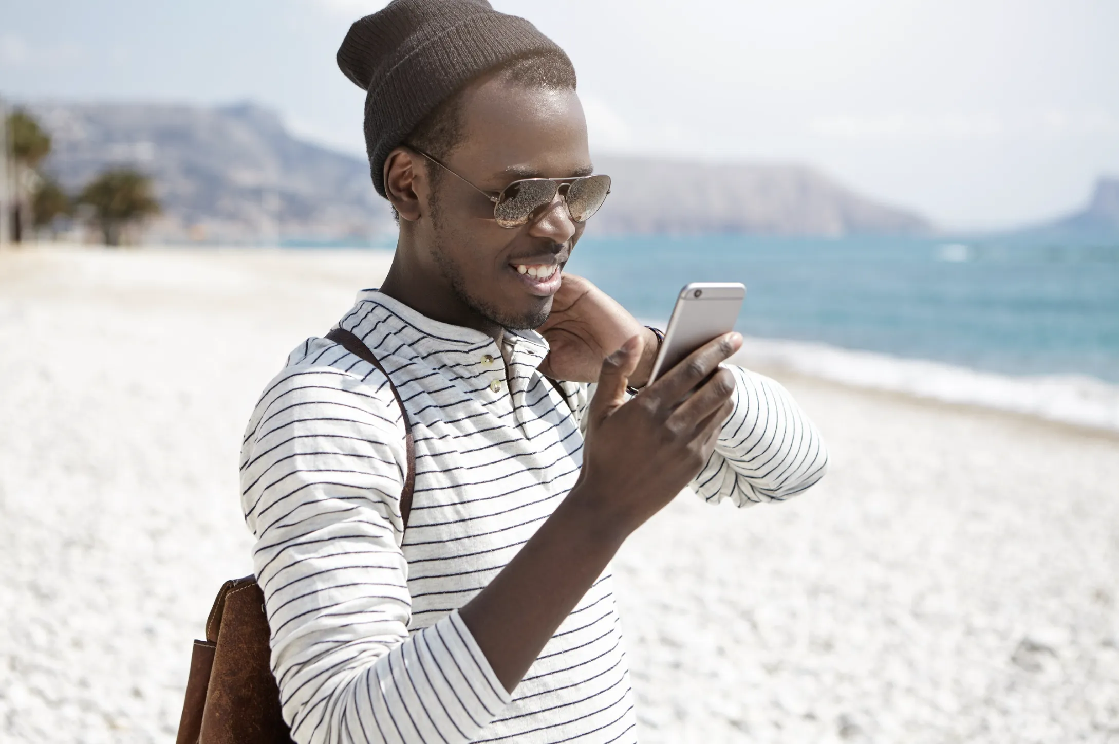 Young man in a beanie and sunglasses smiling at his smartphone on a sunny beach with mountains in the background.