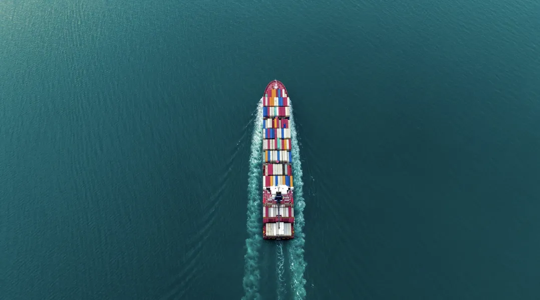 Aerial view of a large cargo container ship sailing through calm teal ocean waters, leaving a white wake trail.