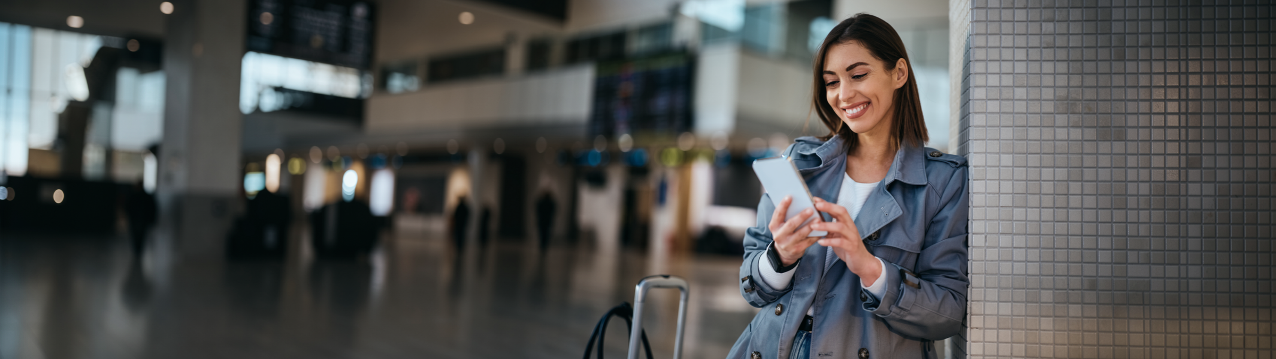 Smiling woman in a trench coat using a smartphone at an airport terminal, leaning against a tiled wall with luggage nearby.