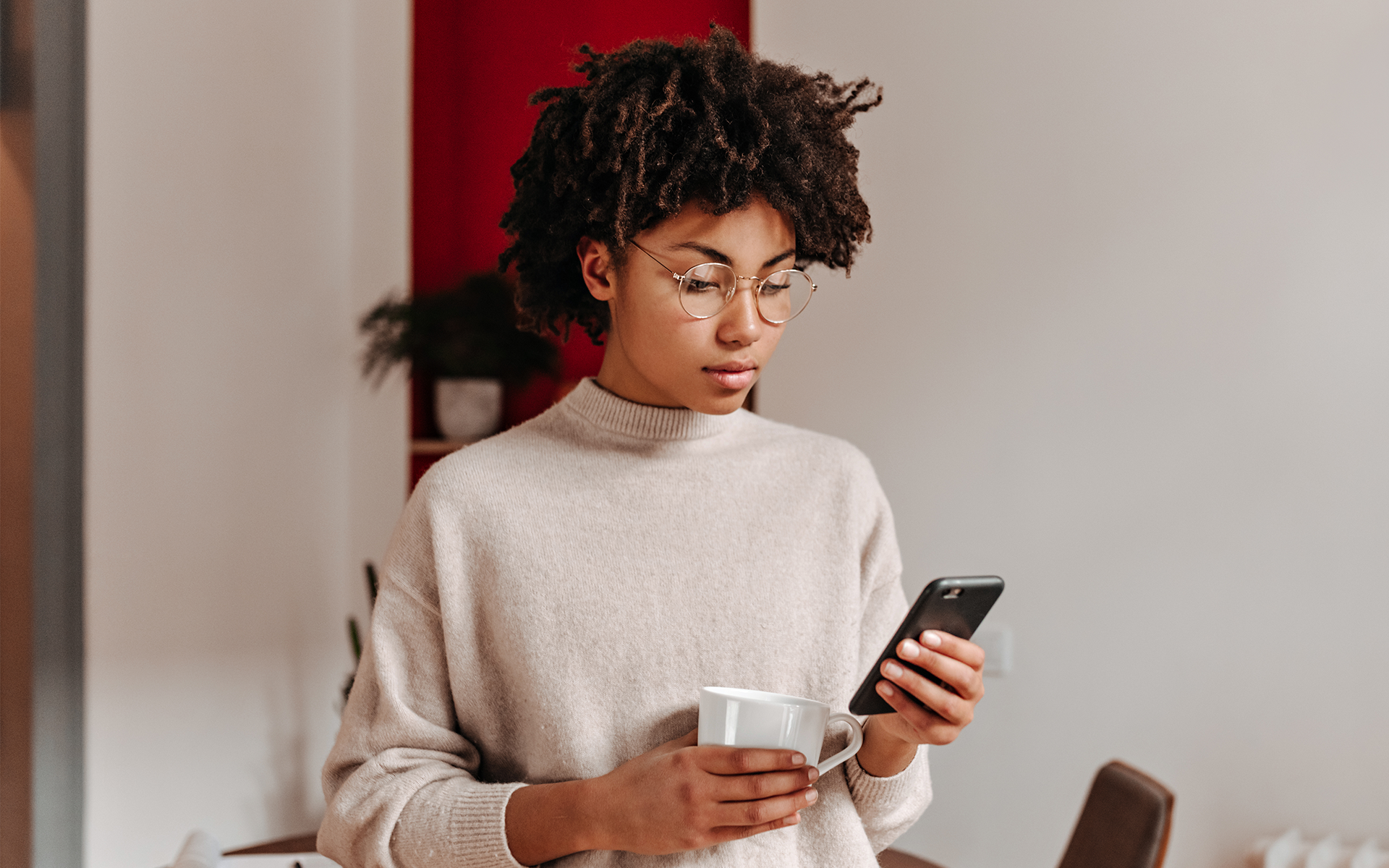 Young woman with curly hair and glasses reading her smartphone while holding a coffee mug indoors.