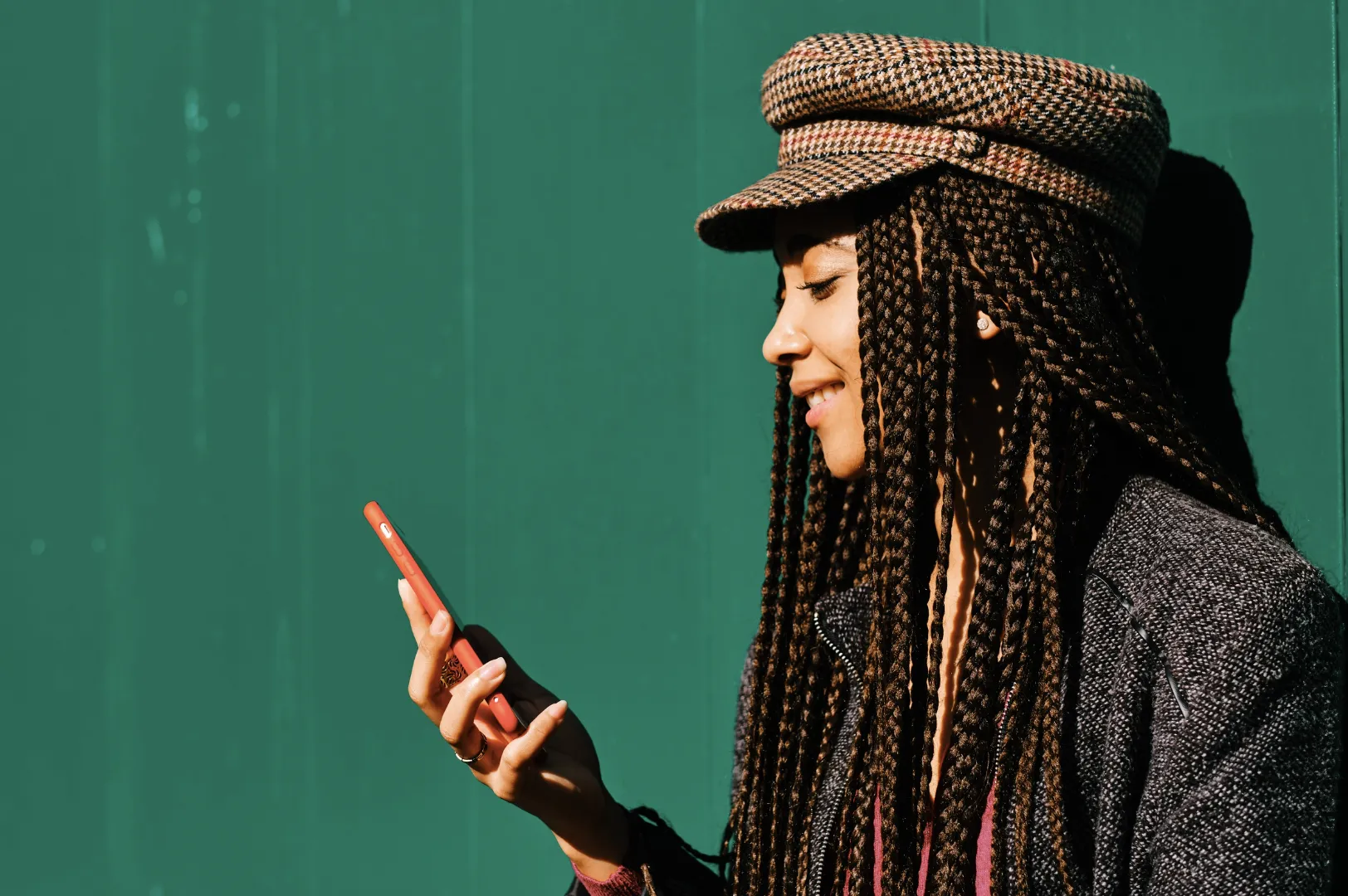 Young woman with long box braids and a tweed flat cap smiling at her smartphone against a teal green wall.