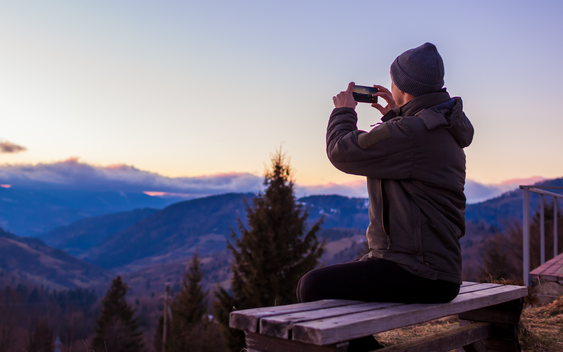 Person in a winter jacket and beanie sitting on a bench, photographing a mountain sunset landscape with a smartphone.