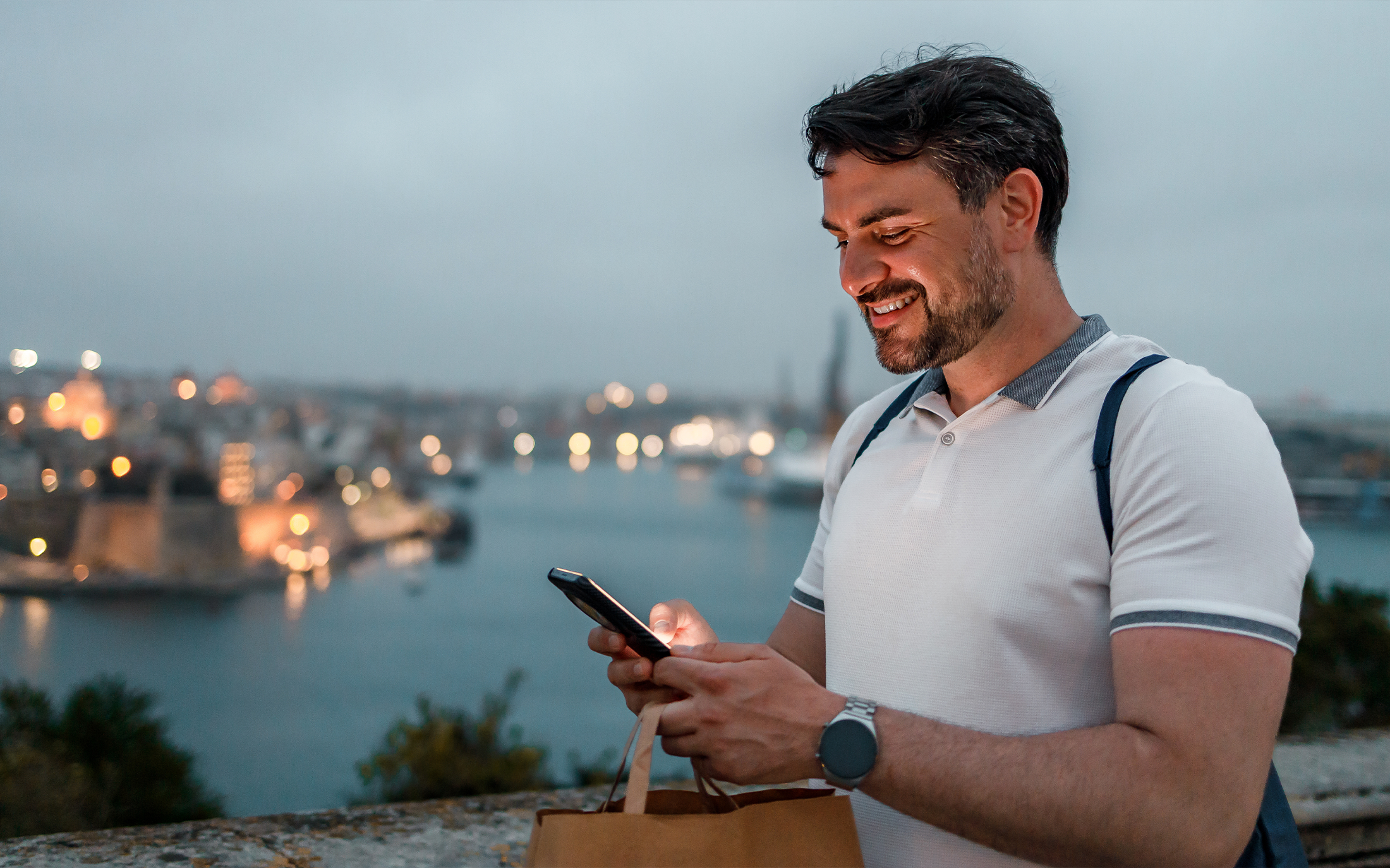 Smiling man using a smartphone outdoors at dusk, holding a shopping bag, with a lit coastal cityscape in the background.