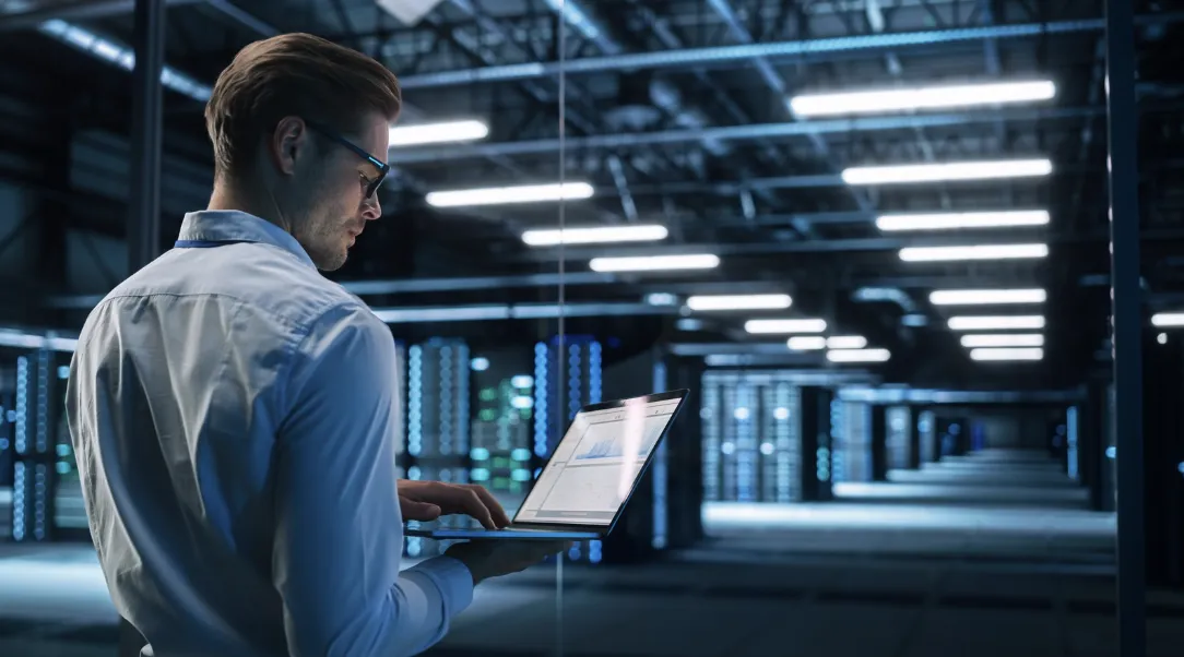IT professional using a laptop in a dimly lit data center with rows of illuminated server racks in the background.