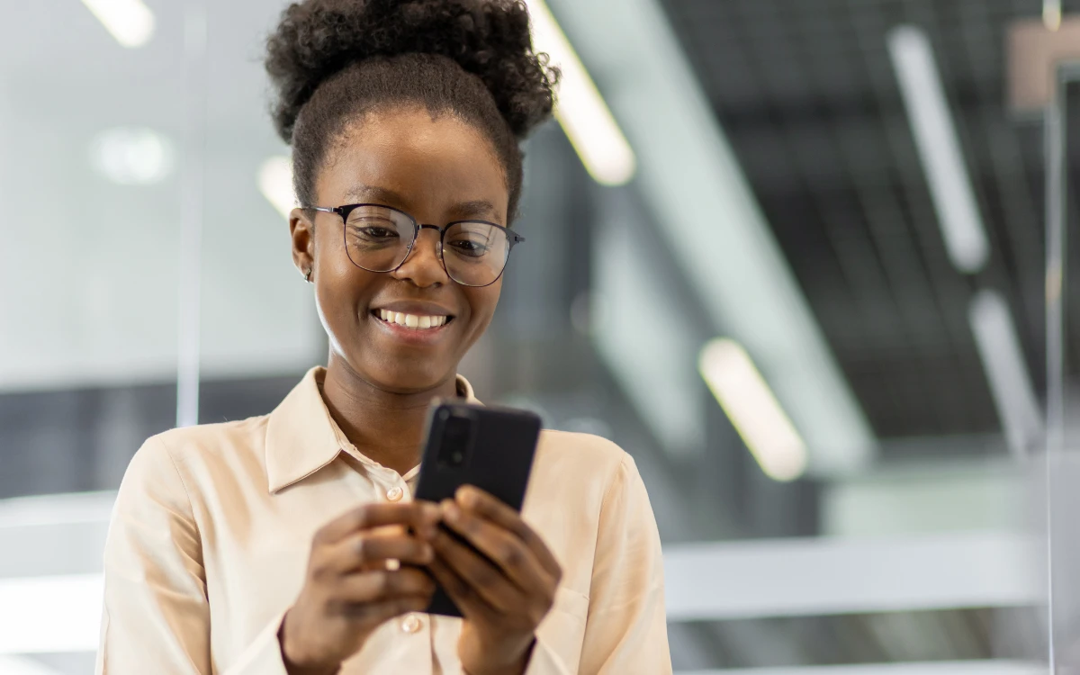 Young woman with glasses smiling while using a smartphone in a modern office setting.