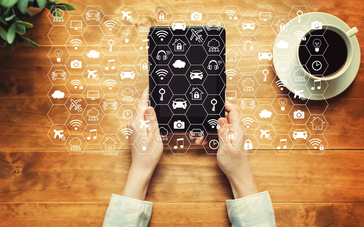 Hands holding a tablet on a wooden desk surrounded by IoT hexagonal icons representing smart devices, Wi-Fi, cars, and cloud connectivity.