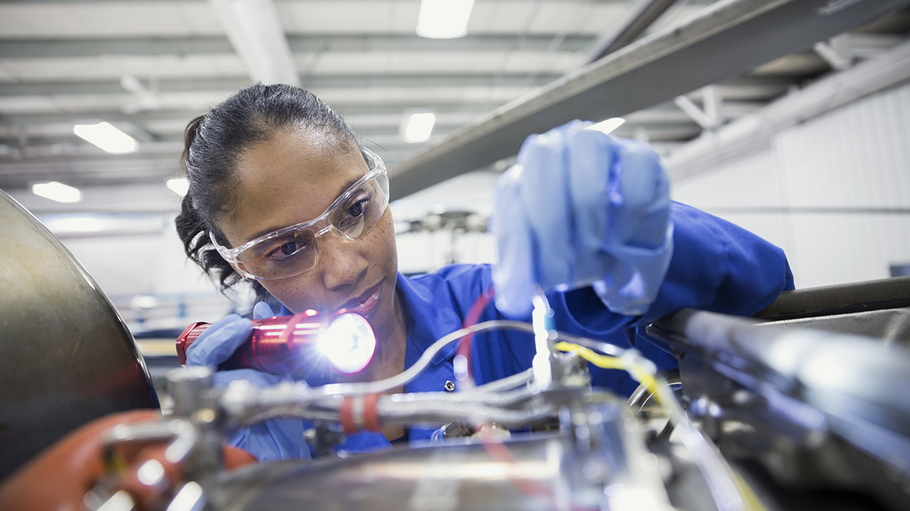 Female mechanic wearing safety goggles and blue gloves inspects a piece of machinery with a flashlight.