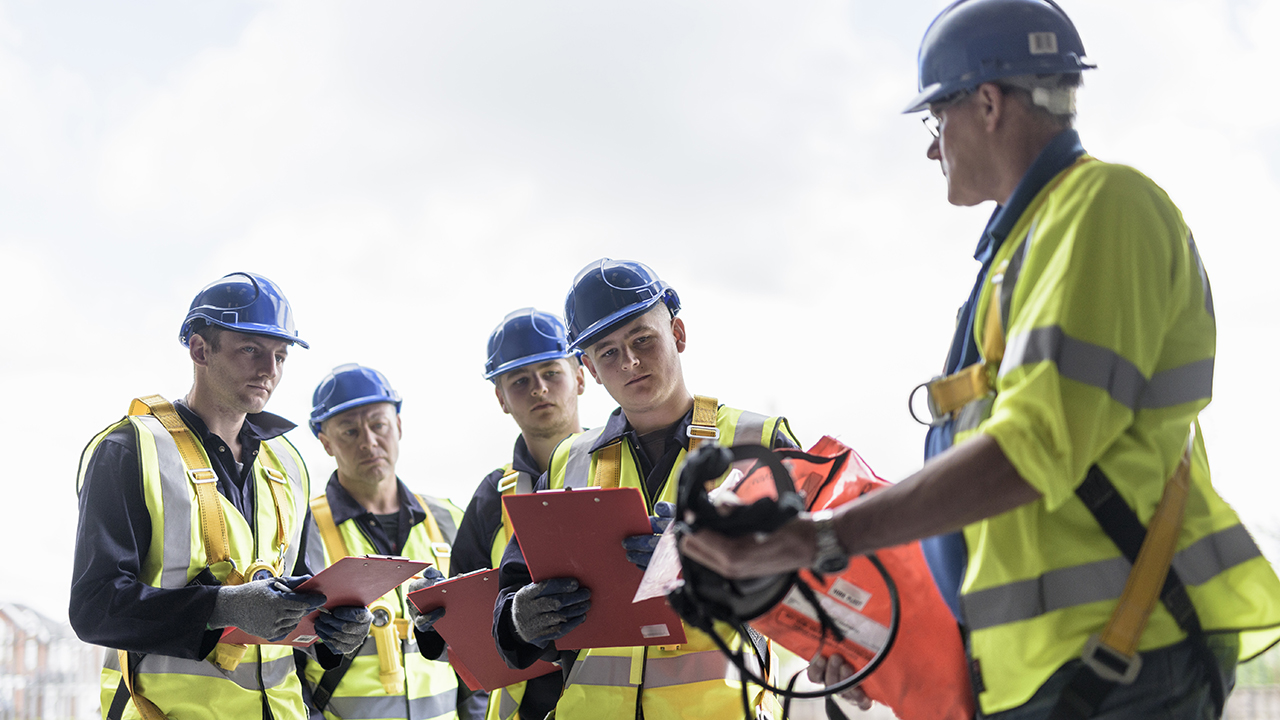 Construction supervisor in yellow safety vest instructs four workers in blue hard hats holding red clipboards outdoors.