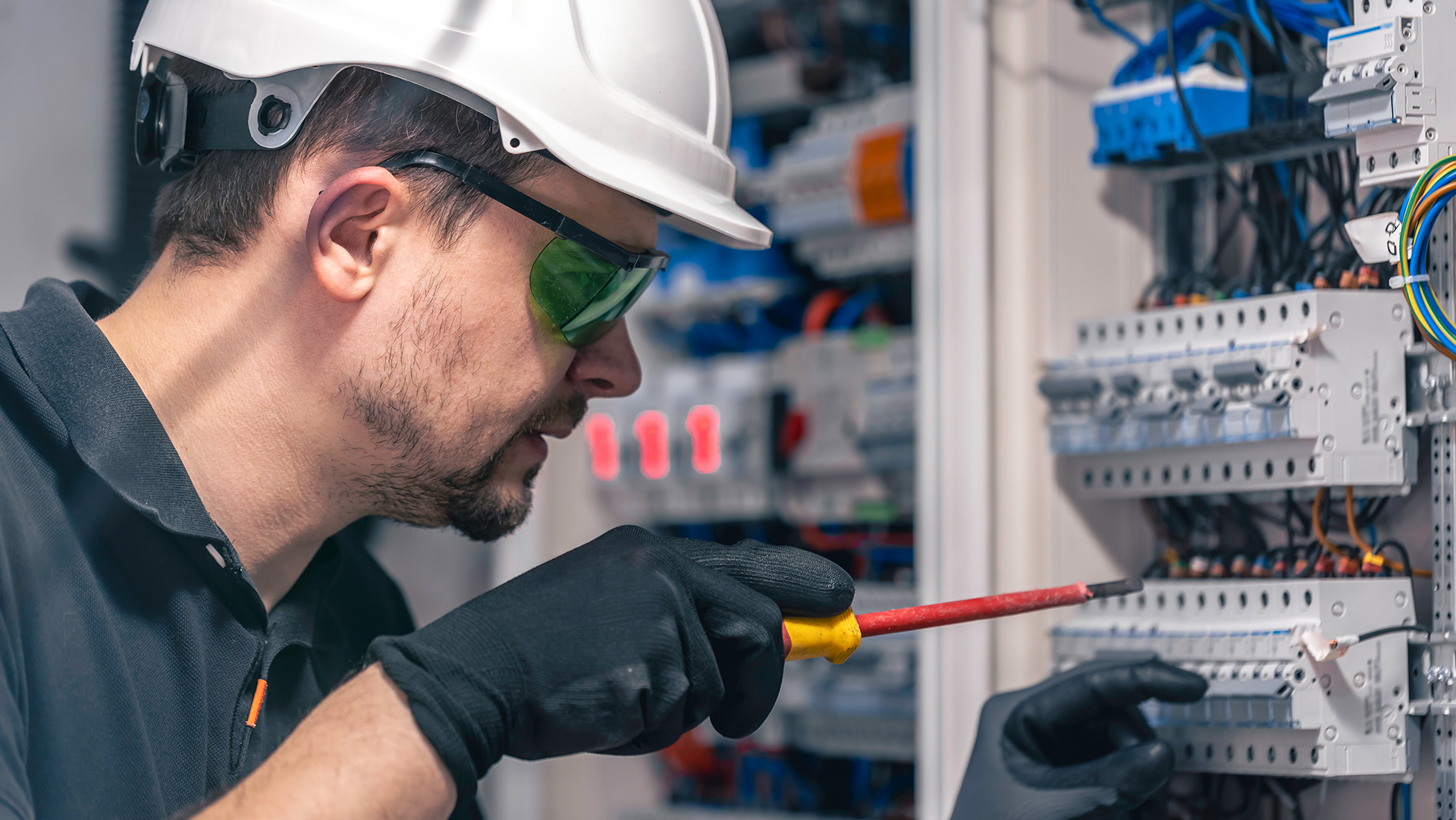 Electrician in white hard hat and safety glasses working on electrical panel with screwdriver in industrial setting.