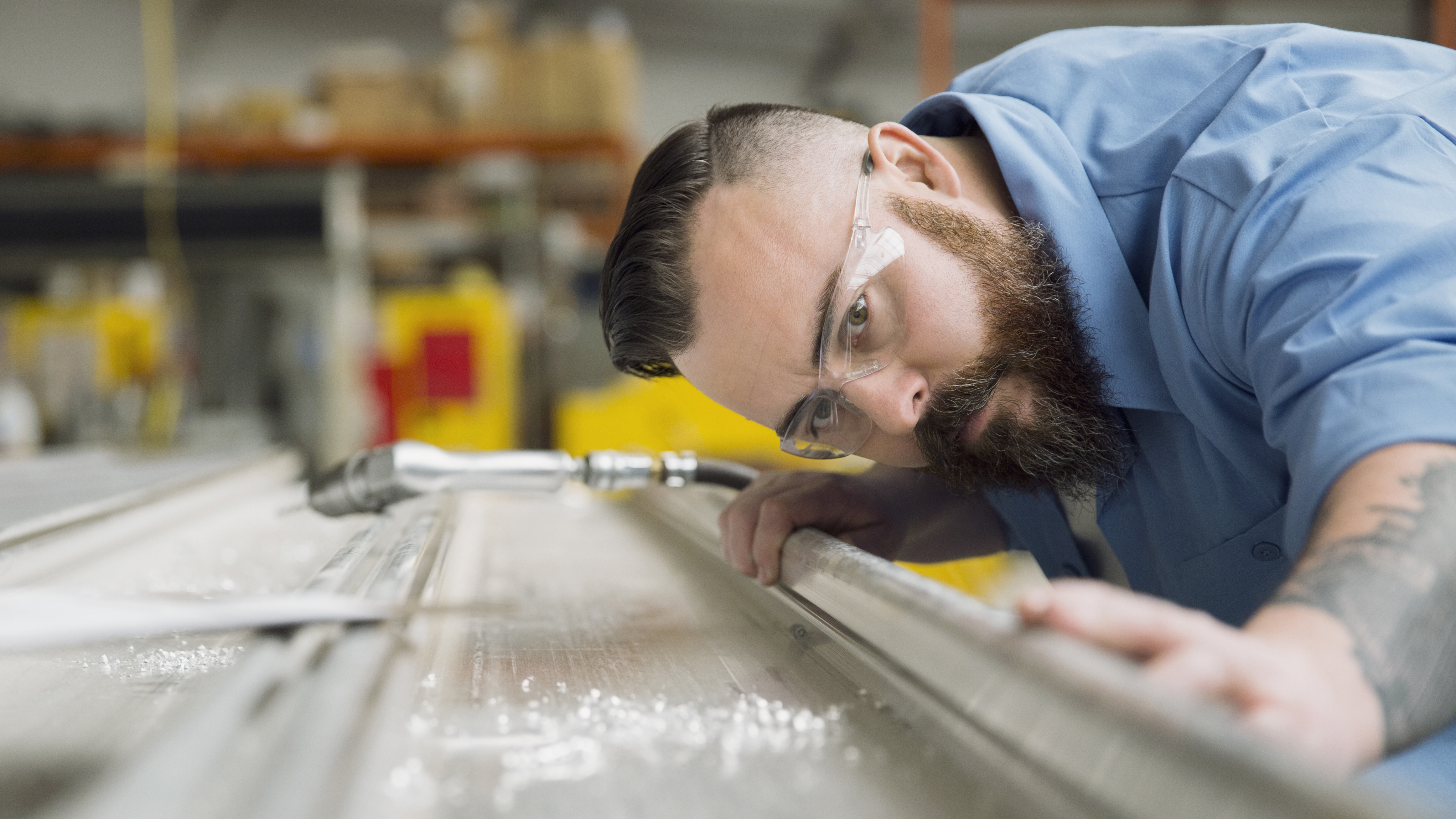 Worker in blue shirt and safety glasses inspecting metal surface in industrial warehouse setting