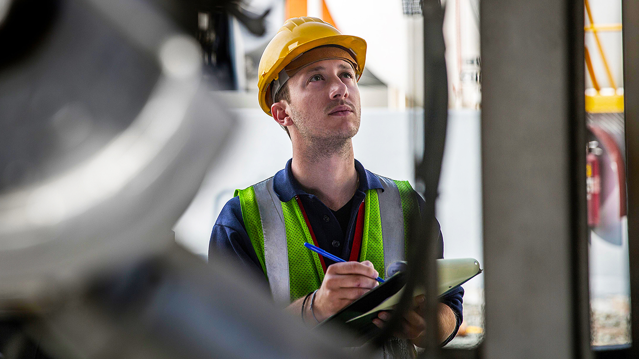 Construction worker in yellow hard hat and green safety vest looking up while holding tablet and pen at worksite.