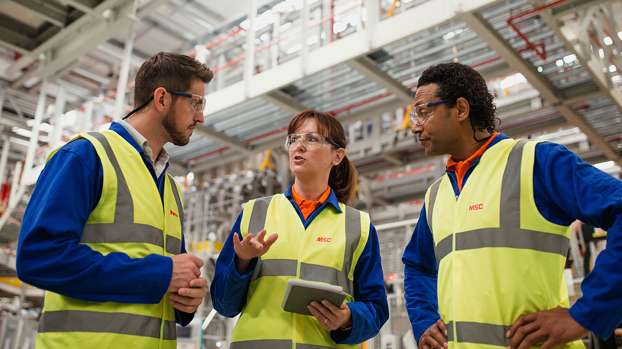 Three warehouse workers in yellow safety vests and blue uniforms with safety glasses discussing work in an industrial facility.