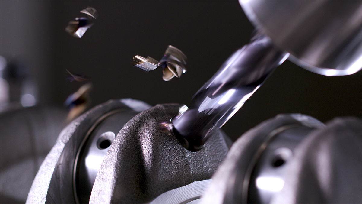 Close-up of a metal drill bit cutting into a dark industrial component, with metal shavings flying off during machining.