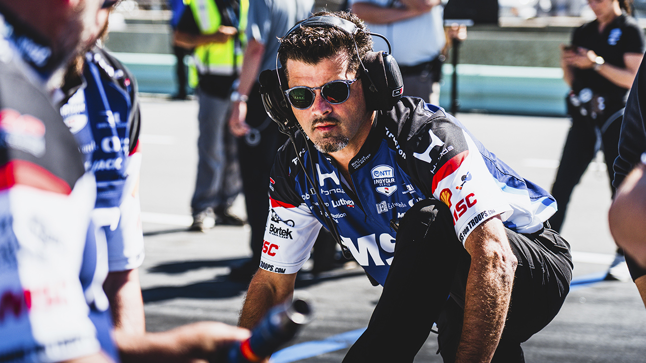 RLL Racing crew member wearing sunglasses and headset crouches in pit lane surrounded by team members in racing uniforms with MSC logo.