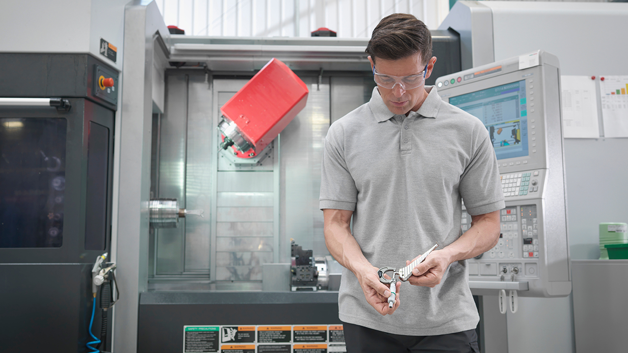 Man in safety glasses examining metal part in front of CNC machine with computer control panel in industrial workshop.