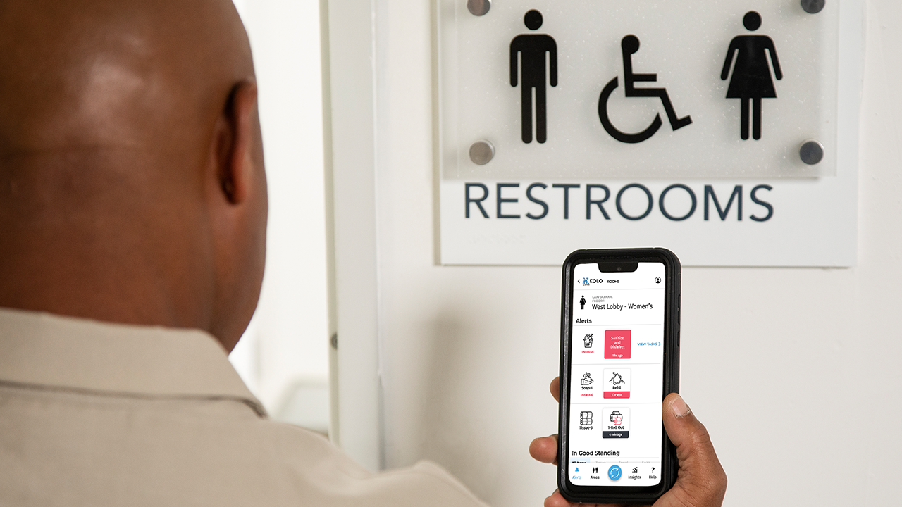 Person holding smartphone displaying restroom facility app in front of accessible restroom sign with gender symbols