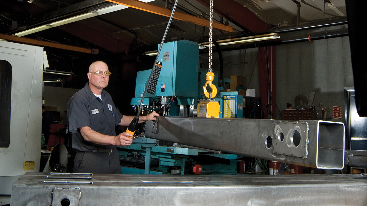 Worker in industrial shop operating yellow chain hoist to lift large metal beam near teal machinery
