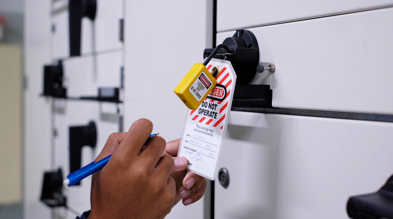 Hand writing on a lockout tagout tag with yellow padlock attached to electrical panel, warning Do Not Operate.