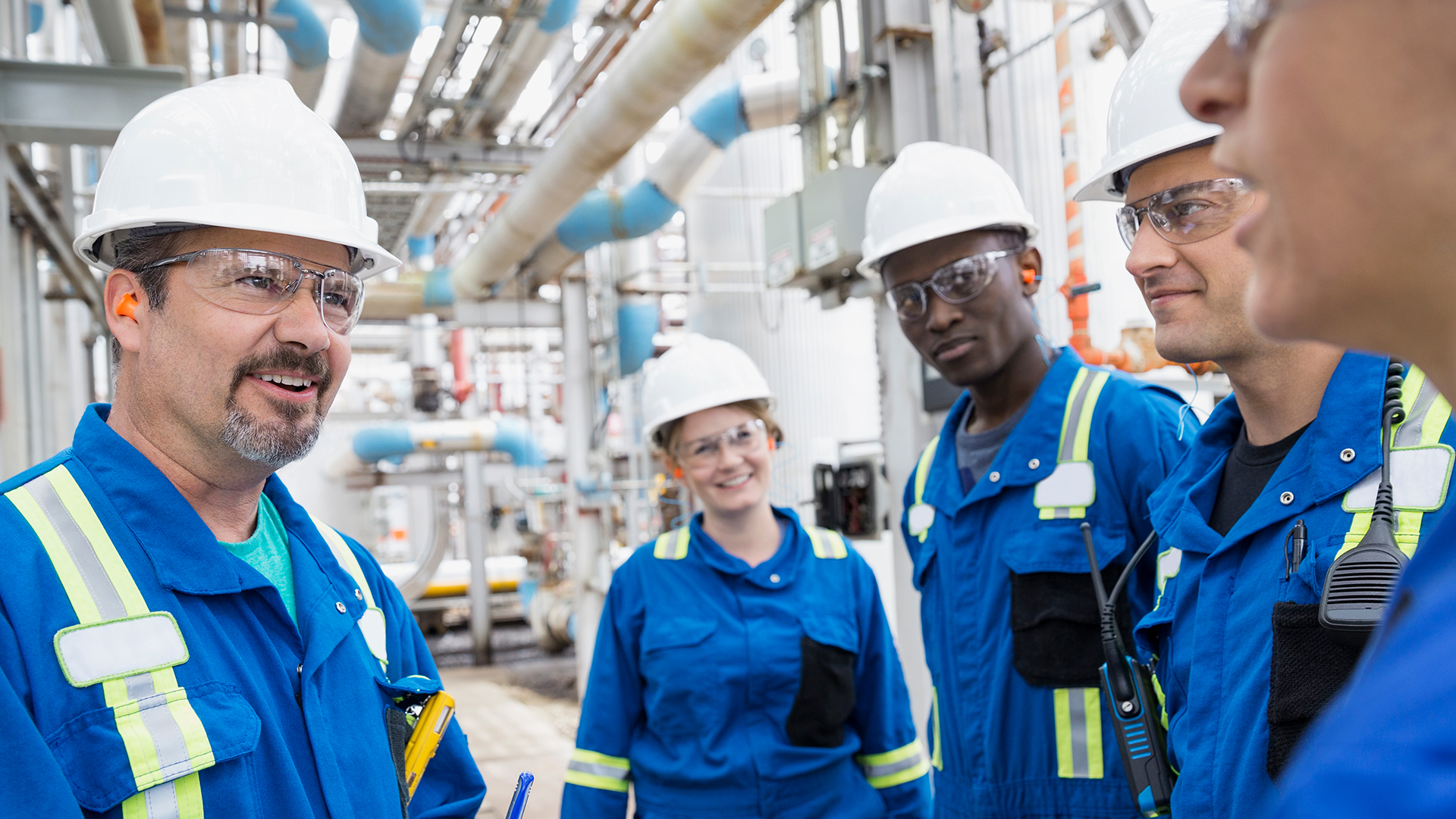 Four industrial workers in blue coveralls, white hard hats, and safety glasses standing in a facility with pipes overhead.