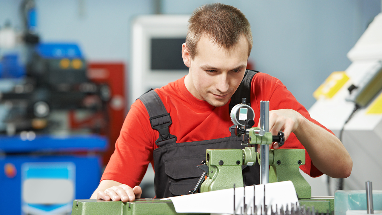 Technician in red shirt and black apron operating green precision measuring equipment in industrial workshop setting.