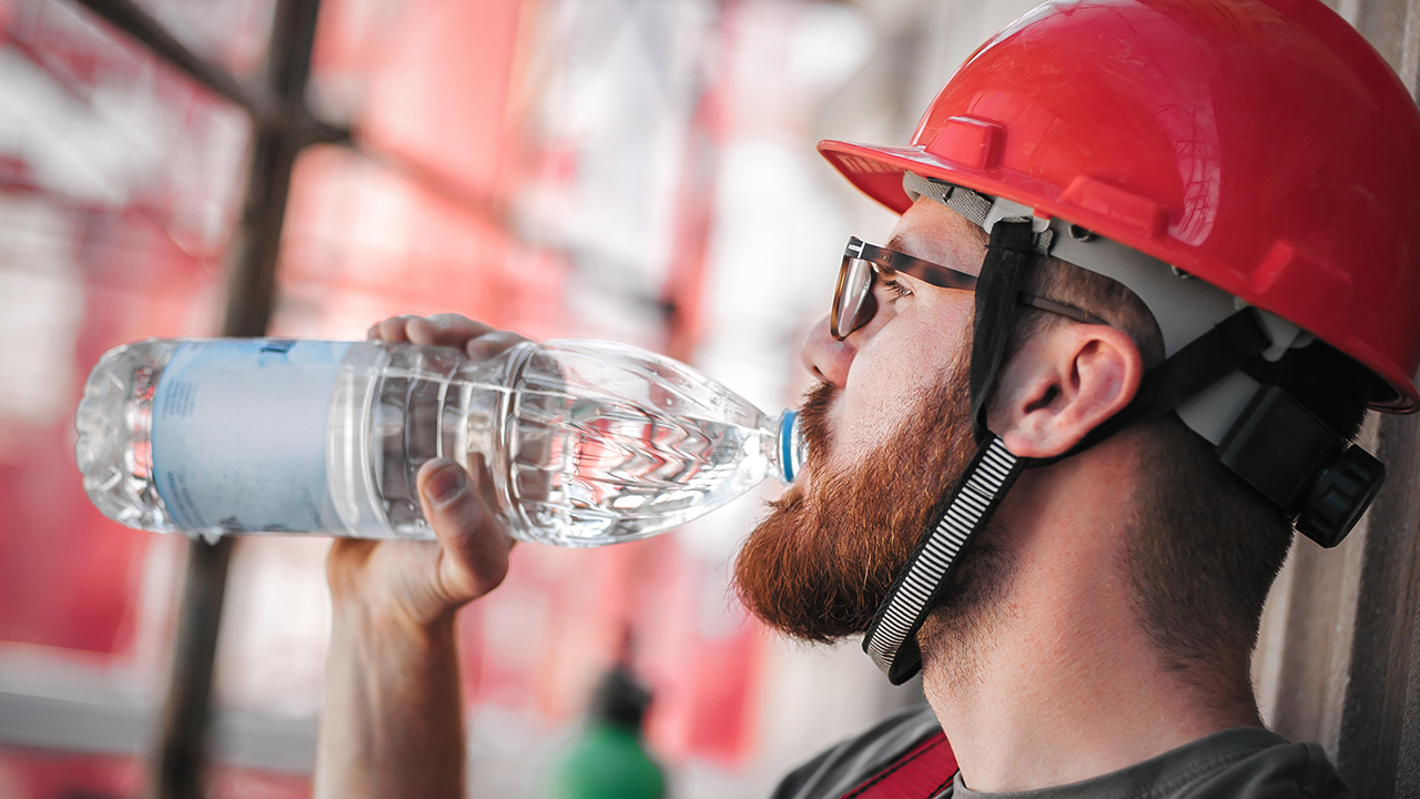 Construction worker with red hard hat and safety goggles drinking water from a clear plastic bottle on site.