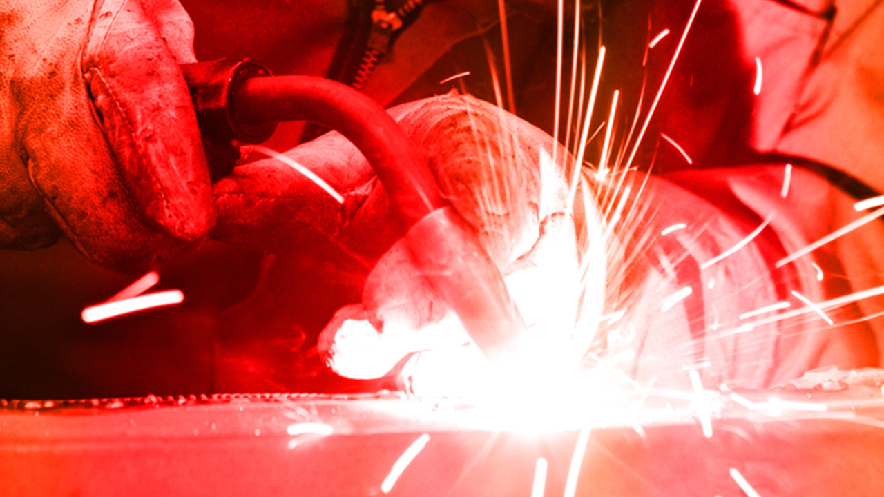 Worker in protective gear using angle grinder on metal surface creating bright white sparks in red-tinted lighting