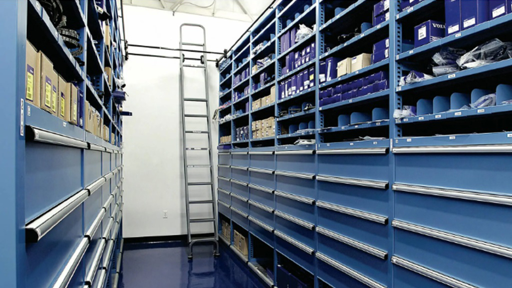Industrial warehouse aisle with blue metal shelving units stocked with parts and supplies, tall ladder in center.