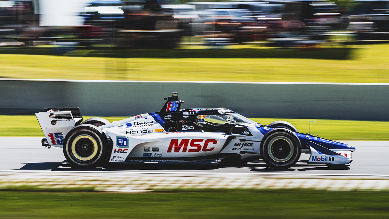 White and blue MSC-branded IndyCar racing on track with motion blur, number 15, Honda powered, speeding past green grass.