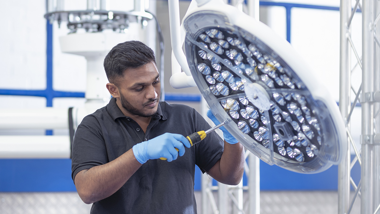 Technician wearing blue gloves using screwdriver to service a large overhead surgical light in medical facility