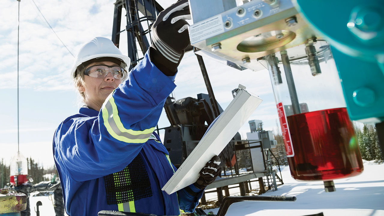 Industrial worker wearing a blue safety jacket and white hard hat inspects oilfield equipment outdoors, holding a metal clipboard near machinery with a red fluid container on a snowy site.