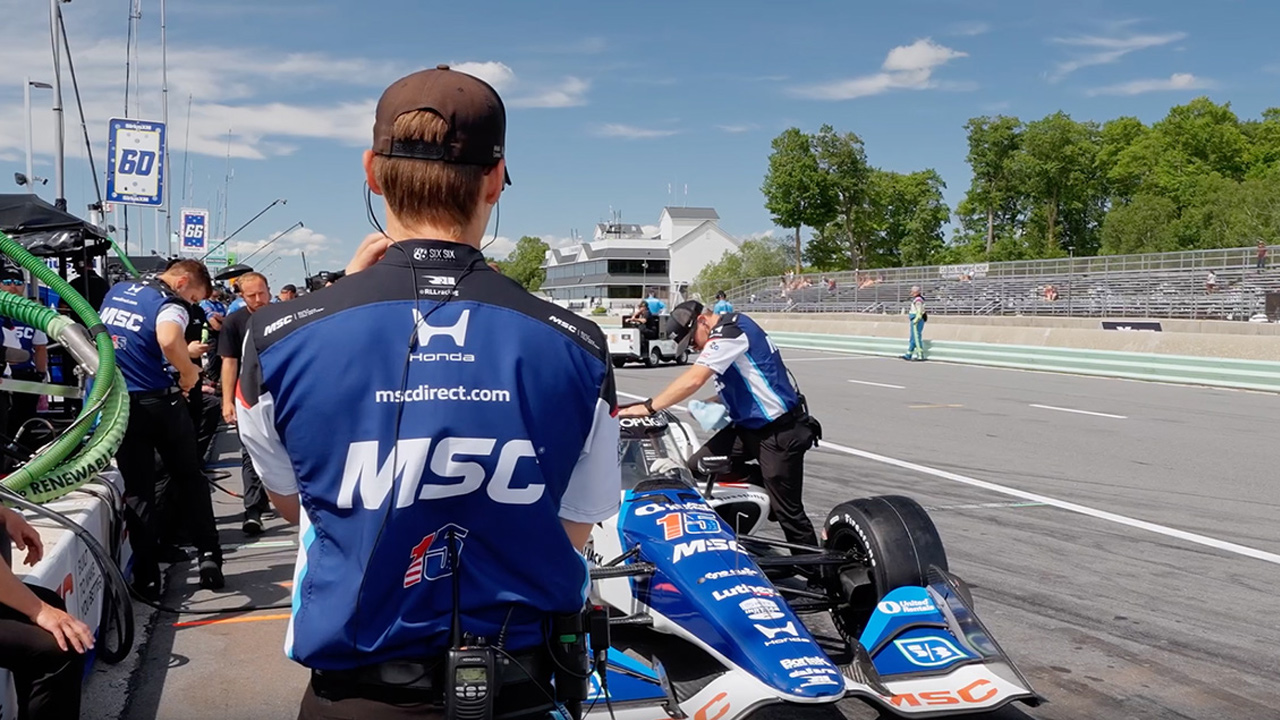 RLL Racing crew member in blue and black racing vest with mscdirect.com branding and MSC logo standing beside race car on pit lane at track