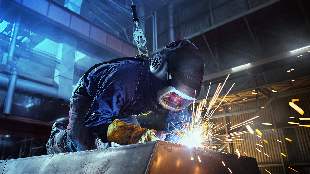 Welder in protective clothing, helmet and gloves welding metal in industrial facility with bright sparks flying.