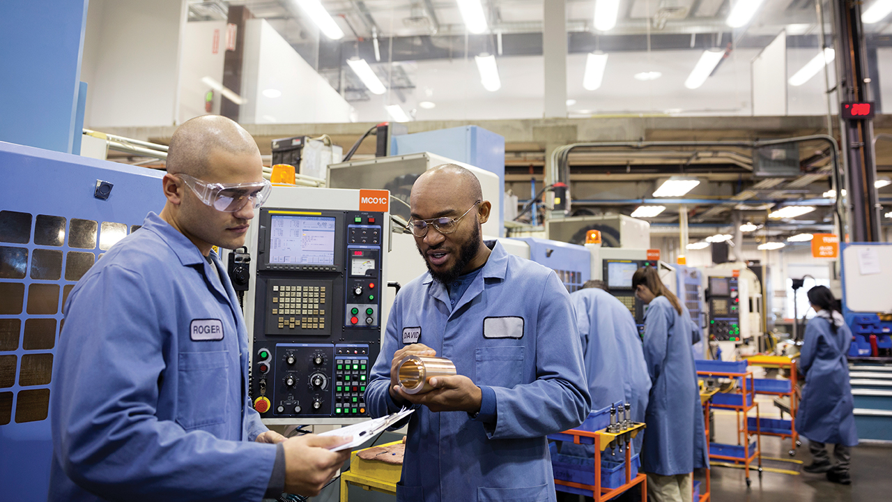 Two workers in blue uniforms inspecting a metal part in front of industrial CNC machinery in a manufacturing facility