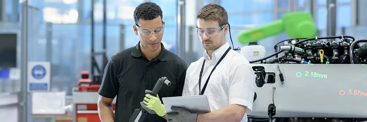 Two technicians wearing safety glasses review a tablet in a modern manufacturing facility with industrial equipment.