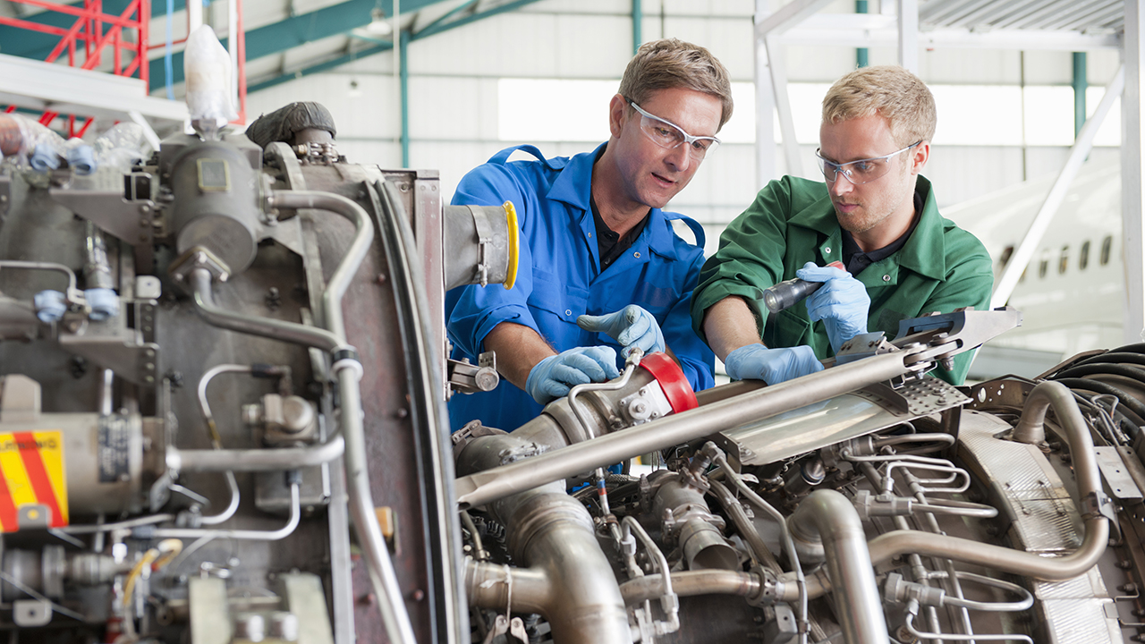 Two technicians in blue and green uniforms wearing safety glasses work together on industrial machinery in a factory setting.