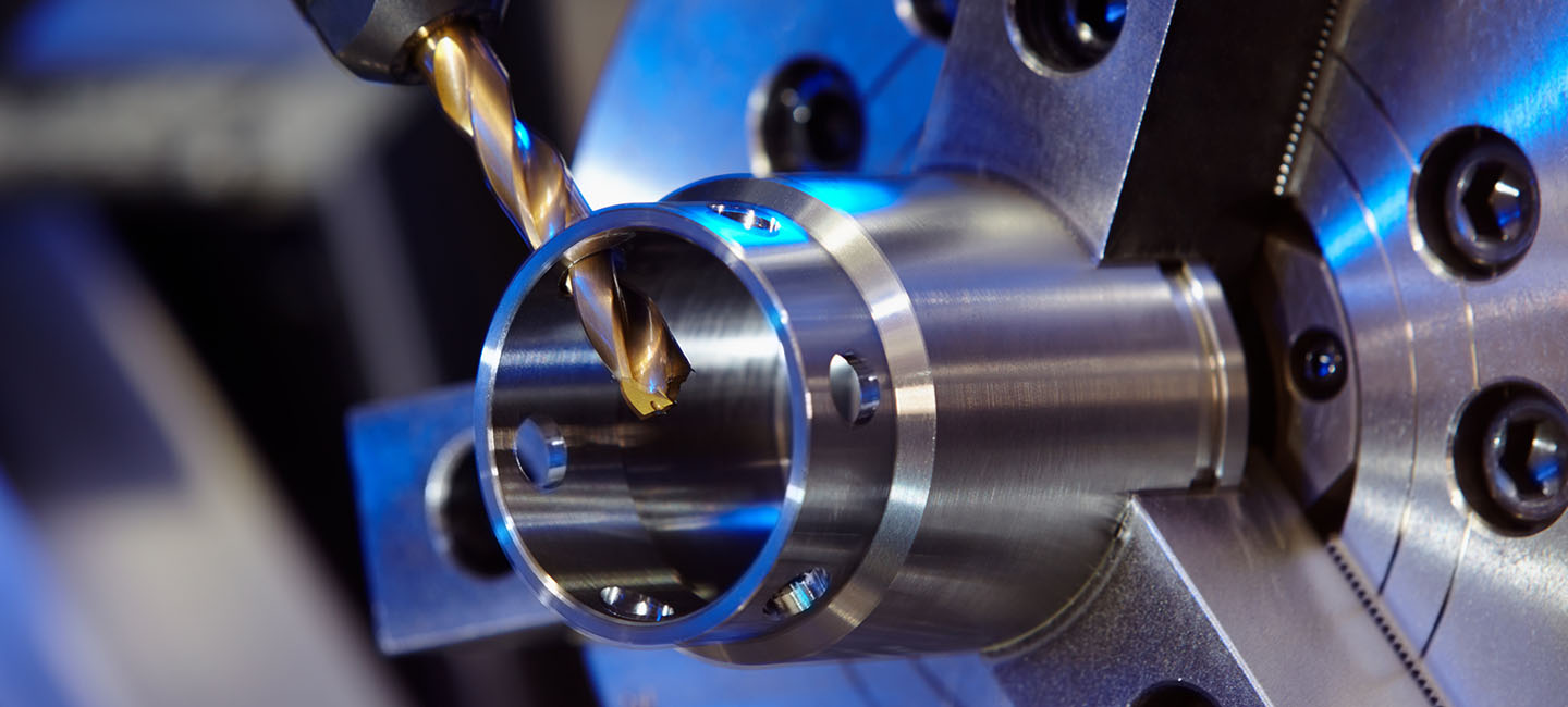 Close-up of a gold drill bit machining the interior of a cylindrical metal part with blue industrial lighting.
