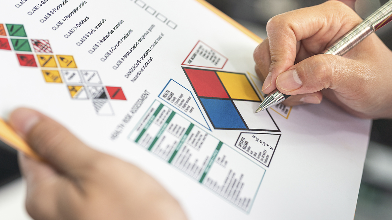 Hand holding pen pointing to HMIS hazard classification chart with colored diamond sections and safety data tables