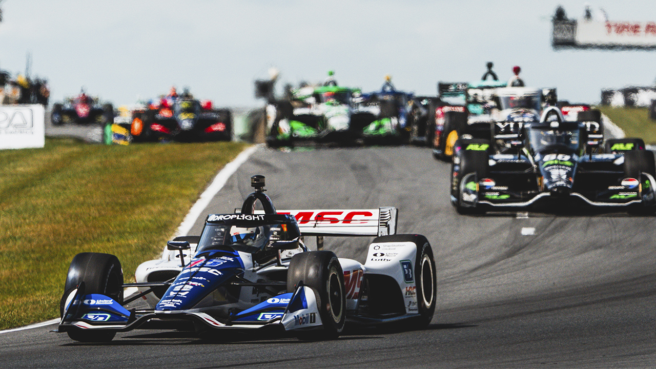 IndyCar race with white and blue car featuring MSC logo on wing, leading pack on track with green grass and overcast sky