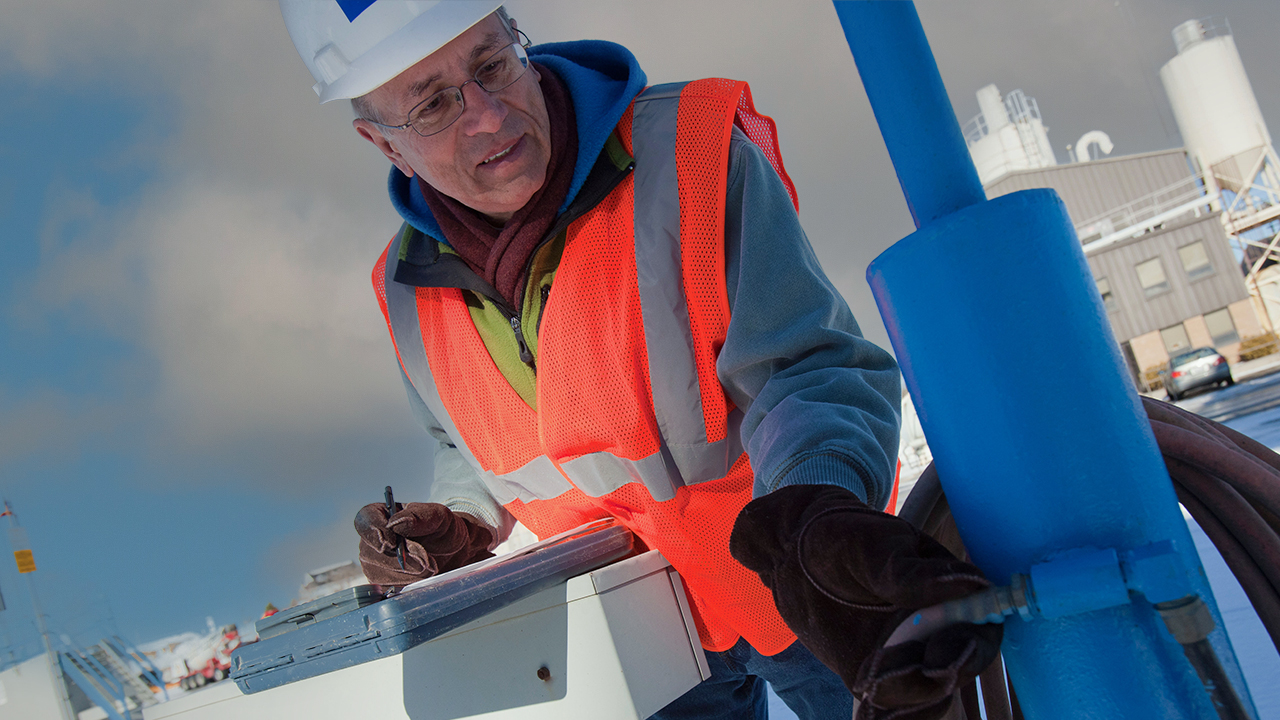 Industrial worker in white hard hat, glasses, and orange safety vest working at industrial facility
