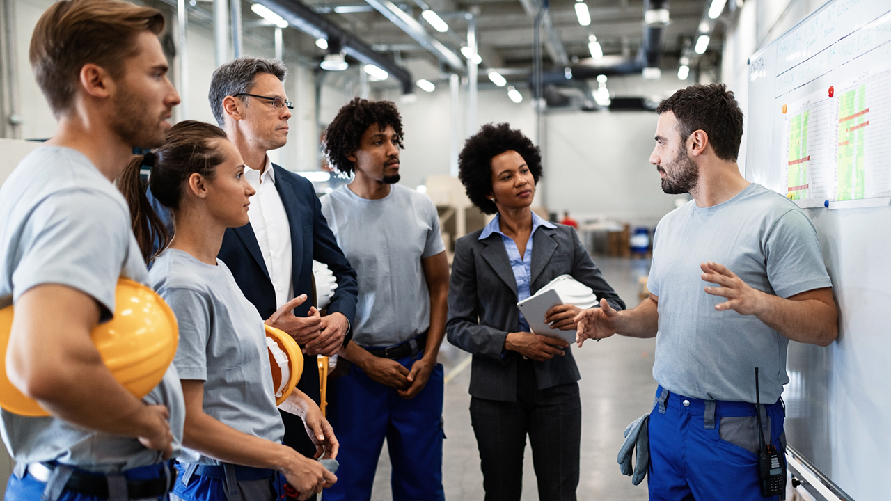 Worker in gray shirt and blue pants gestures while explaining to diverse group of colleagues in industrial facility.